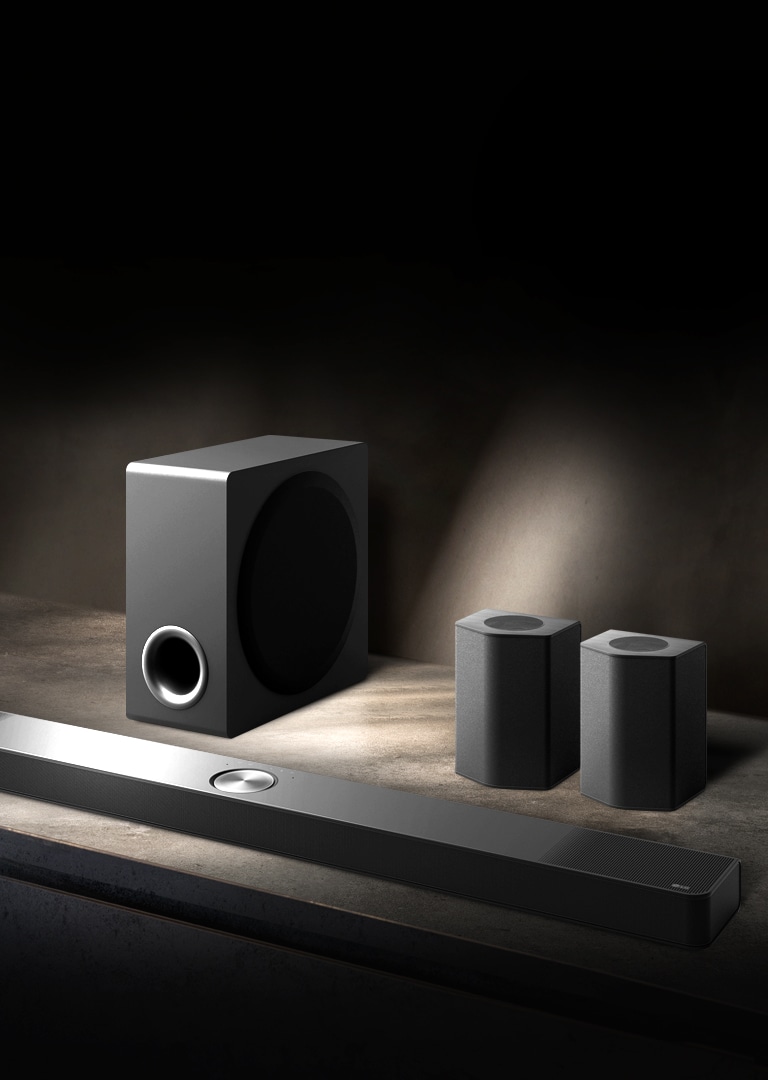 LG Soundbar, enceintes arrière et caisson de basse installés en biais sur une étagère en bois marron dans une pièce sombre, avec une lumière qui n’éclaire que le système audio.