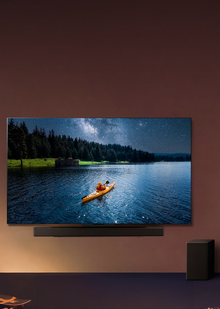 LG TV et Soundbar montés sur un mur avec un caisson de basse sur le sol à droite. La TV affiche une personne en train de faire du kayak sur un lac, tandis que des ombres couvrent le mur.