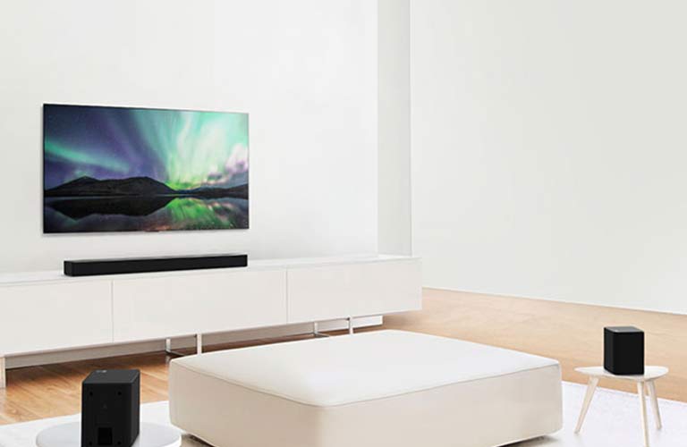 Téléviseur et barre de son dans le salon blanc avec un canapé blanc au centre. Les haut-parleurs sont placés de part et d’autre du canapé.