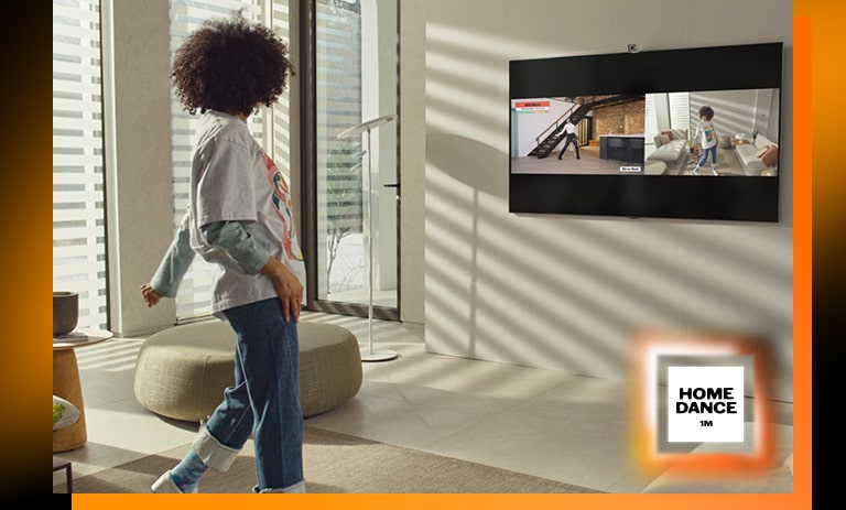 Une femme se tient debout au milieu du salon, danse et regarde la télévision en même temps. Le téléviseur montre une scène de didacticiel de danse sur la gauche et reflète une femme qui danse sur le côté droit de l’image. Le logo « 1million home dance » est visible dans le coin inférieur droit.