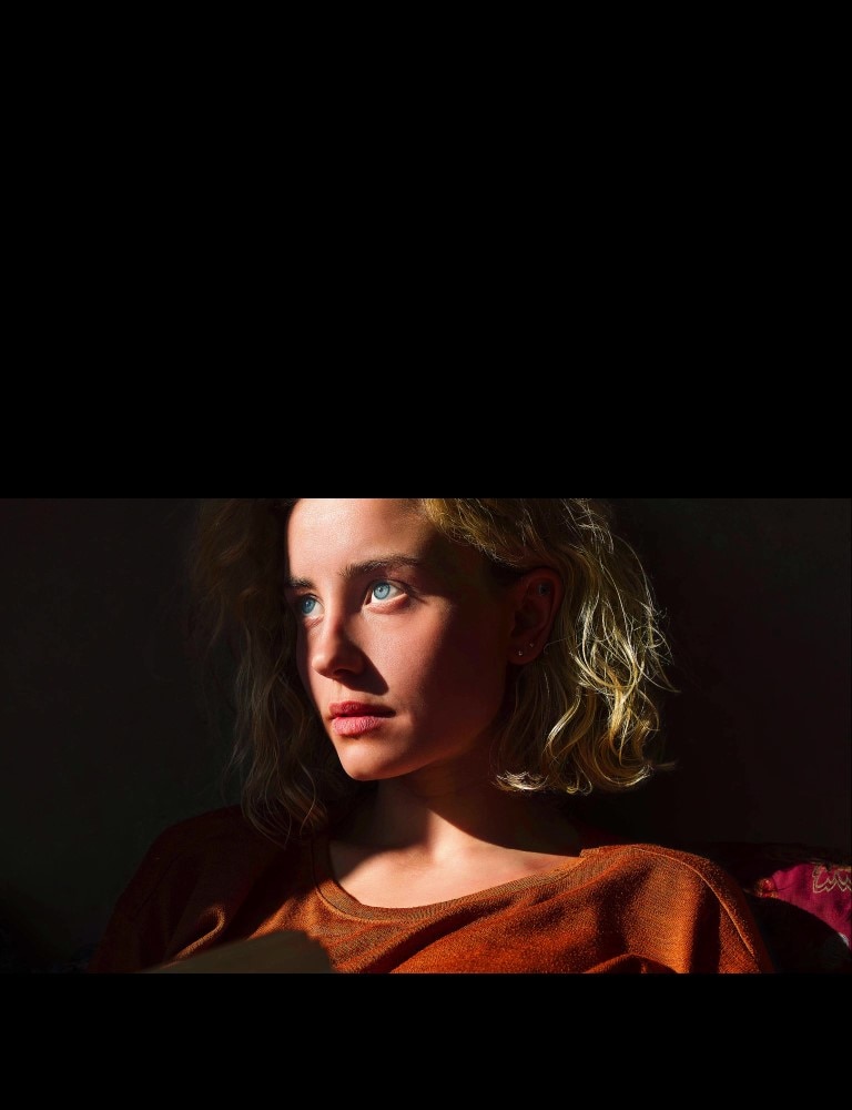 Une femme avec des yeux bleus perçants et un top orange brûlé dans un espace sombre. Des lignes rouges représentant les ajustements IA couvrent une partie de son visage qui est clair et riche en détail, tandis que le reste de l’image semble terne.