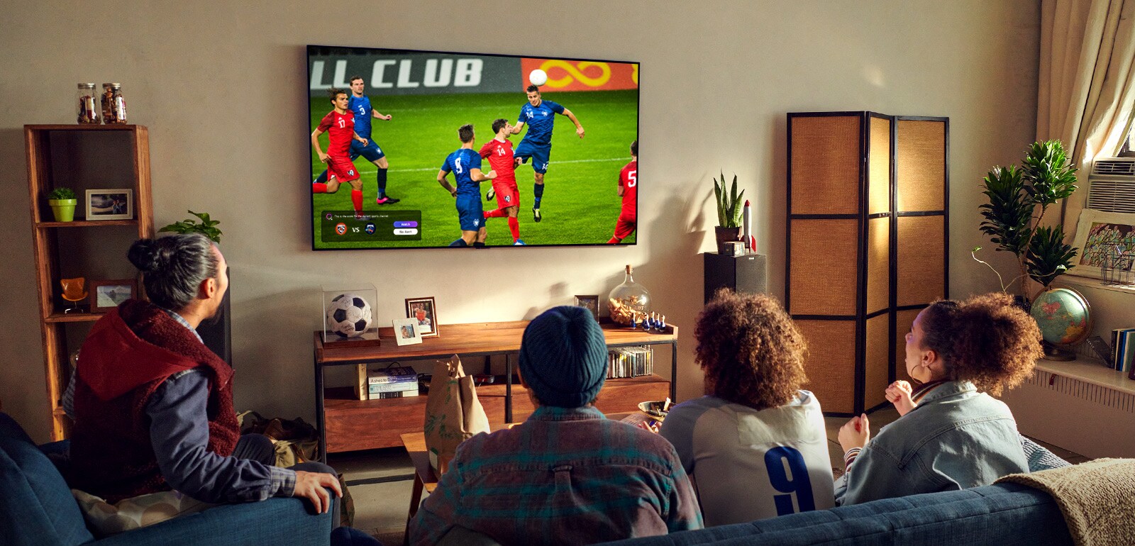 Un homme et une femme assis derrière une table basse et devant un téléviseur fixé au mur qui diffuse un match de football.