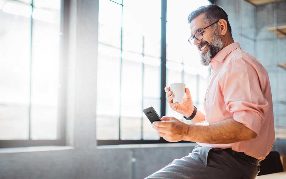 A man holding onto a mug whilst smiling at his LG Smartphone. 