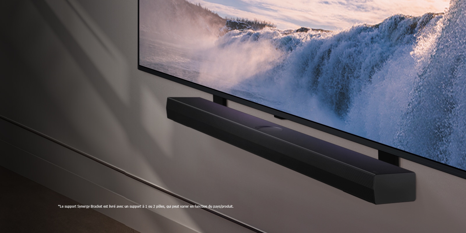 Vue inclinée en perspective d’un LG TV et d’une Soundbar montés sur un mur. La TV affiche un gros plan d’une cascade imposante, alors que la lumière du soleil illumine le mur, la TV et la Soundbar. Une notice indique : « Le support Synergy Bracket est livré avec un support à 1 ou 2 pôles, qui peut varier en fonction du pays/produit. »
