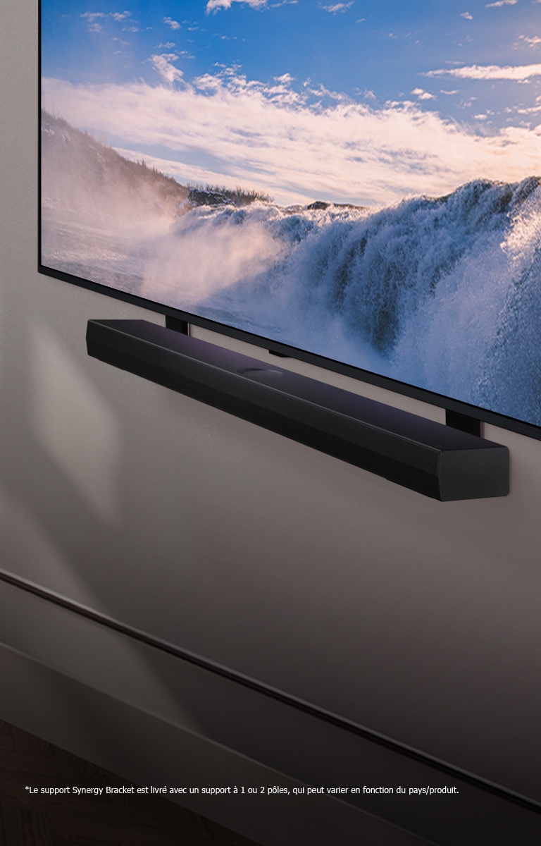 Vue inclinée en perspective d’un LG TV et d’une Soundbar montés sur un mur. La TV affiche un gros plan d’une cascade imposante, alors que la lumière du soleil illumine le mur, la TV et la Soundbar. Une notice indique : « Le support Synergy Bracket est livré avec un support à 1 ou 2 pôles, qui peut varier en fonction du pays/produit. »