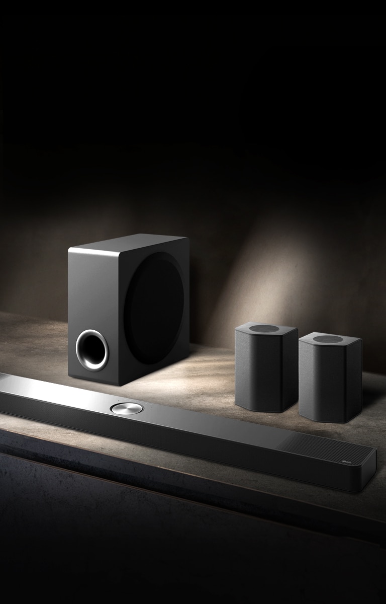 LG Soundbar, enceintes arrière et caisson de basse installés en biais sur une étagère en bois marron dans une pièce sombre, avec une lumière qui n’éclaire que le système audio.