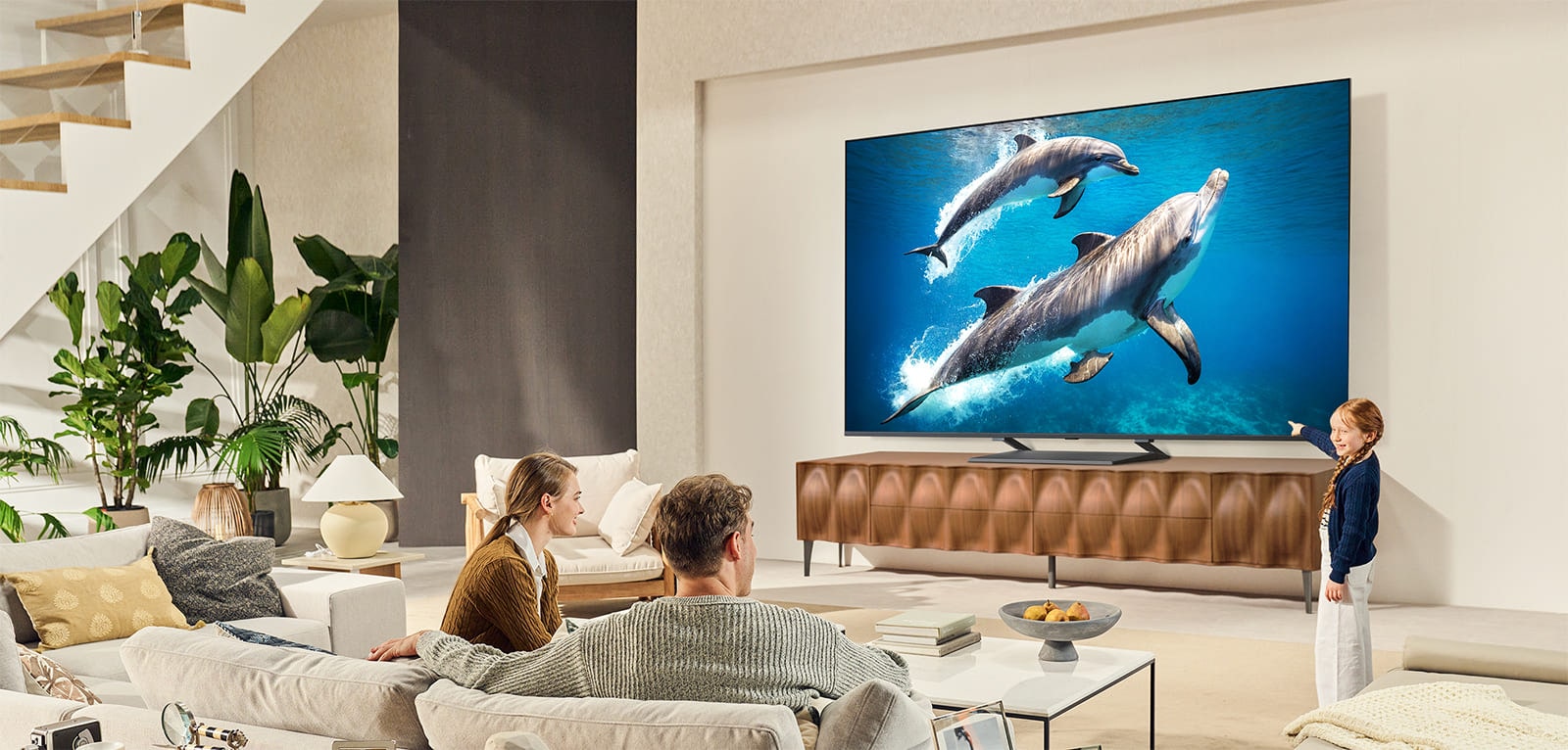 Семейство седи на диван с лице към LG QNED TV, монтиран на стената над LG Саундбар, а малко момиченце сочи към екран, на който се показват два делфина. 