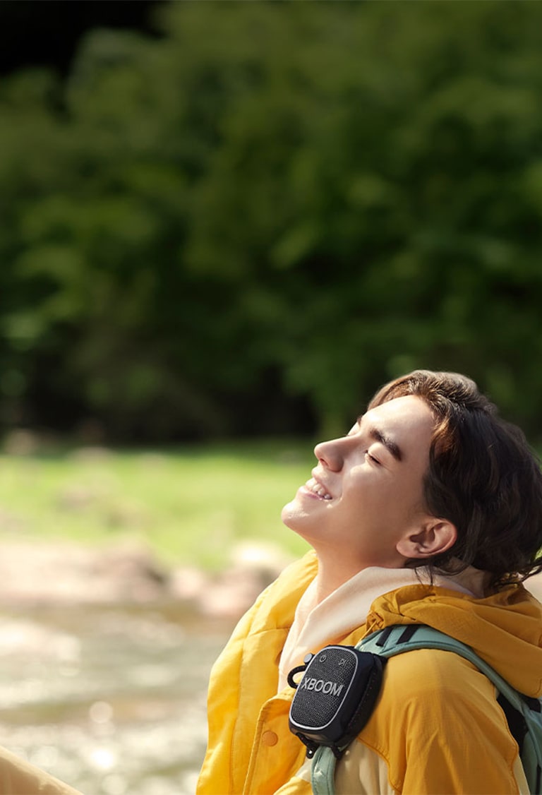 Човек, който седи на пода и слуша музика с LG XBOOM Go XG2T, закачен на раницата му.