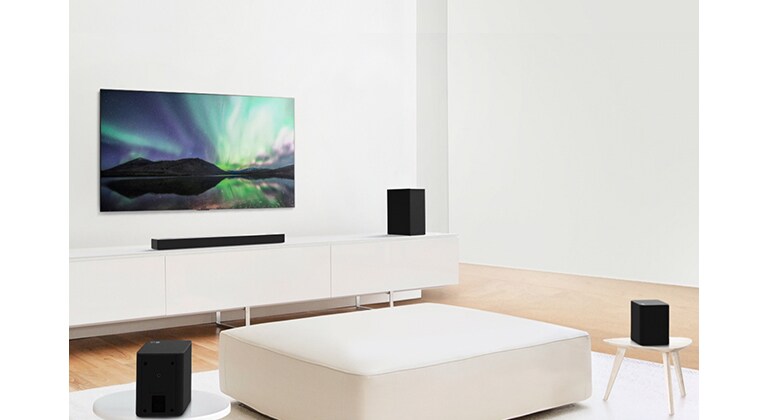 Телевизор и саундбар в бяла всекидневна, в центъра на която има бял диван. Високоговорителите са поставени в двата края на дивана.