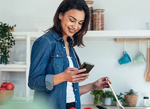 Изображение на жена, която получава известие на смартфона си от приложението LG ThinQ за това, че вратата на хладилника е оставена отворена, докато тя готви.