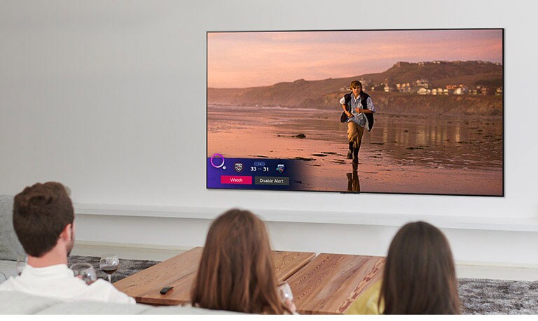 Трима души гледат телевизионен екран, показващ сцена от фентъзи филм с предупреждение за спортно събитие