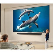 LG 65-инчов LG QNED AI QNED70 4K Smart TV 2025, Семейство седи на диван с лице към LG QNED TV, монтиран на стената над LG Soundbar, а малко момиченце сочи към екран, на който се показват два делфина. Заглавието разказва за това как всяко действие е, 65QNED70A6A, thumbnail 10