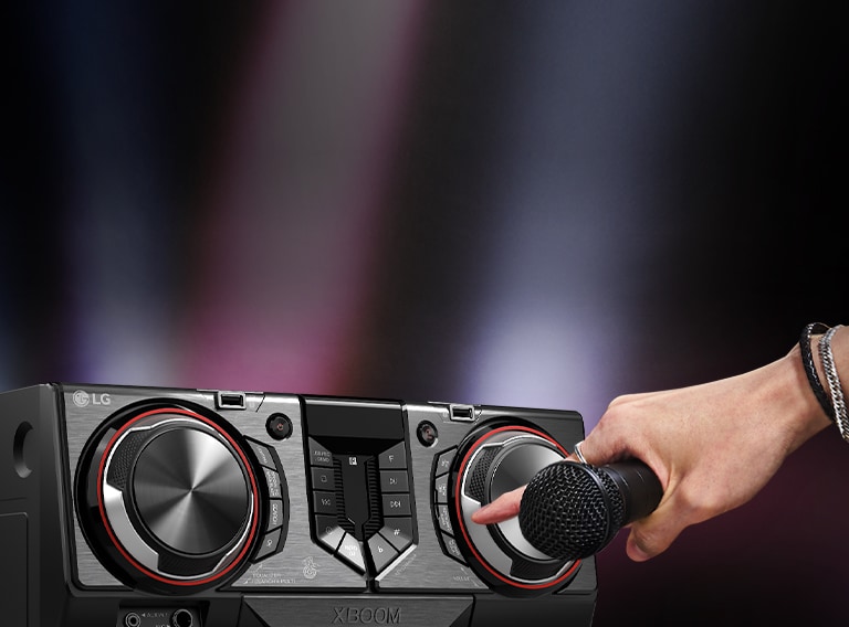 La imagen muestra un fondo borroso. En el lado izquierdo de la imagen, hay un primer plano de la vista lateral del receptor, con una mano sosteniendo un micrófono. El dedo índice se extiende para presionar el botón de karaoke.