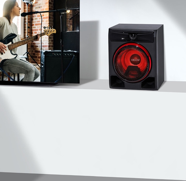 La imagen muestra un primer plano de una larga mesa de televisión blanca. A la izquierda, un televisor LG muestra una escena de un concierto de guitarra. En el lado derecho del televisor, se coloca un altavoz sobre la mesa.