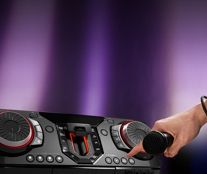 La imagen muestra un fondo borroso con focos de color violeta. En el lado izquierdo de la imagen, hay un primer plano de la vista lateral del receptor, con una mano sosteniendo un micrófono. El dedo índice se extiende para presionar el botón de karaoke.