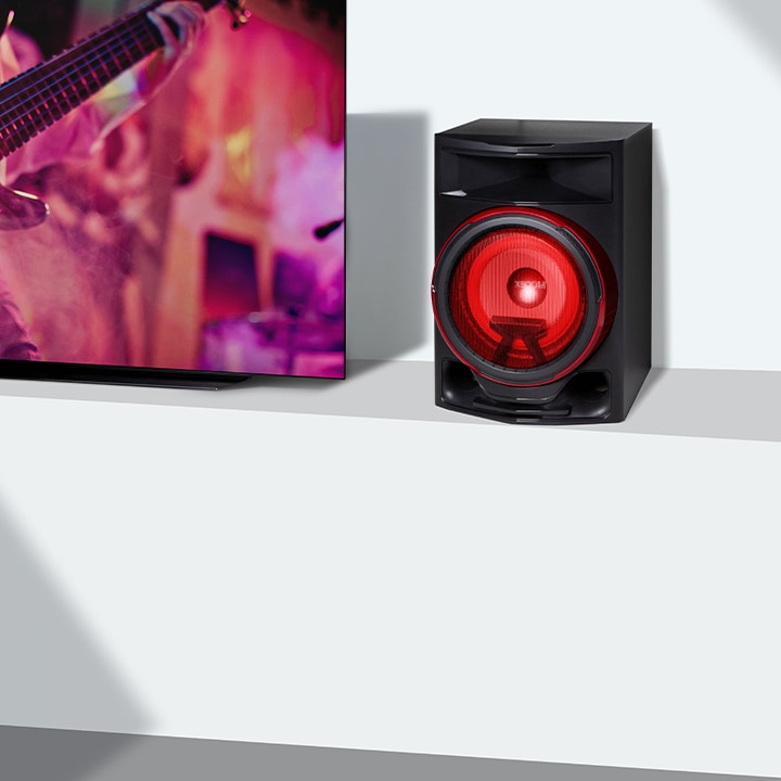 La imagen muestra un primer plano de una larga mesa de televisión blanca. A la izquierda, un televisor LG muestra una escena de un concierto de guitarra. En el lado derecho del televisor, se coloca un altavoz sobre la mesa.