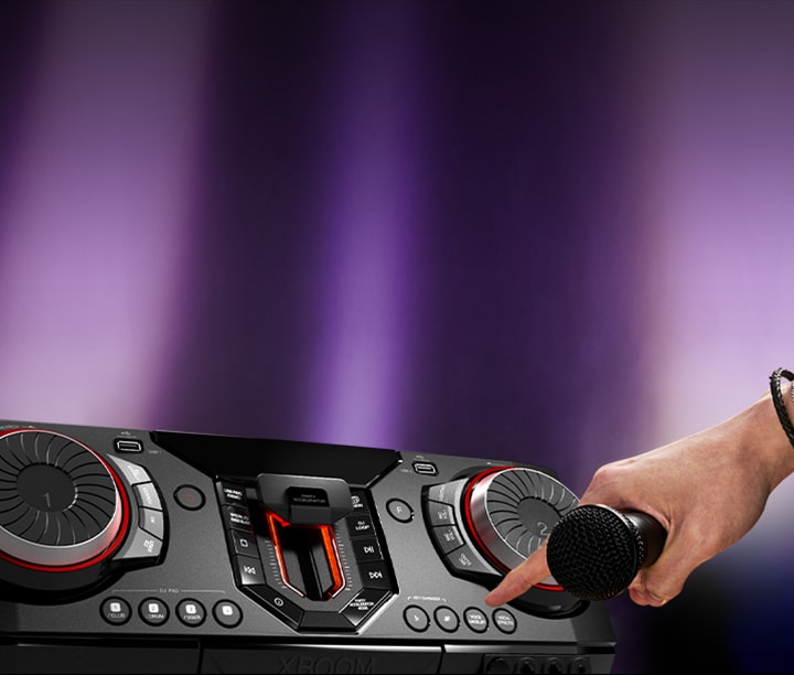 La imagen muestra un fondo borroso con focos de color violeta. En el lado izquierdo de la imagen, hay un primer plano de la vista lateral del receptor, con una mano sosteniendo un micrófono. El dedo índice se extiende para presionar el botón de karaoke.