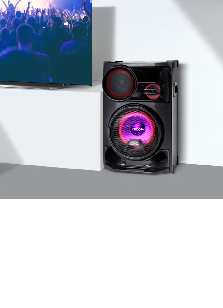 La imagen muestra un primer plano de una larga mesa de televisión blanca. A la izquierda, un televisor LG muestra una escena de concierto. En el lado derecho del televisor, se coloca un altavoz sobre la mesa.