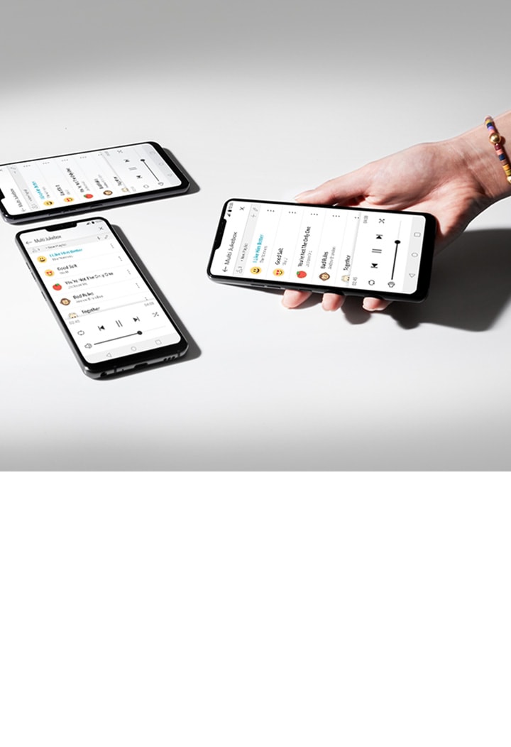 La imagen muestra una superficie blanca con luz proveniente del lado izquierdo. Se colocan tres teléfonos inteligentes sobre la superficie blanca y una mano sostiene uno de ellos. Todos los teléfonos inteligentes muestran la lista de reproducción Multi Jukebox.