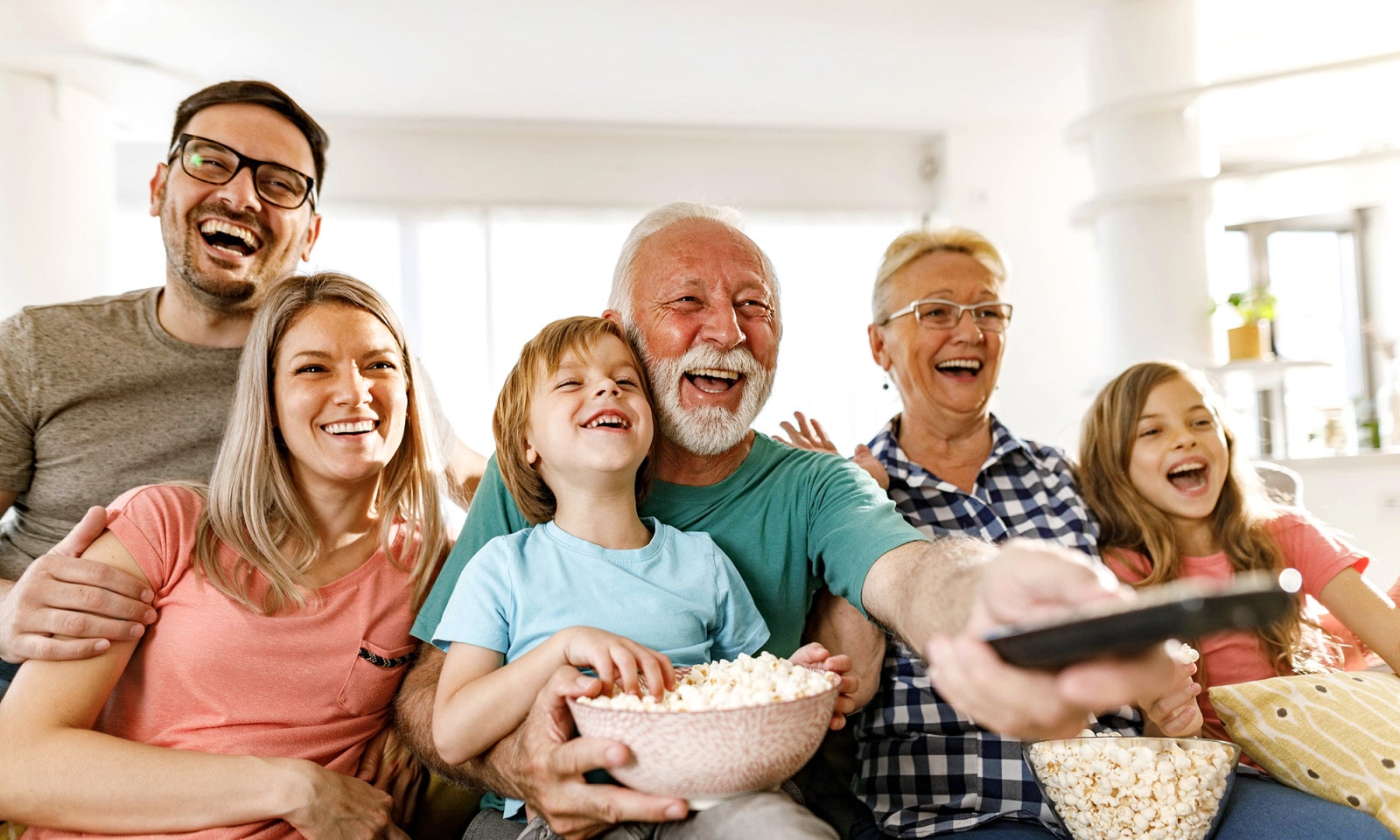 Una familia sentada riendo mientras el abuelo apunta con el control remoto.