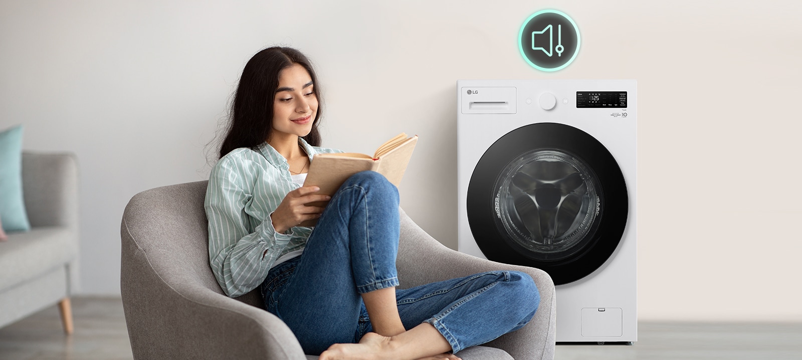 Hay una lavadora en la sala y una mujer está sentada en una silla leyendo un libro.