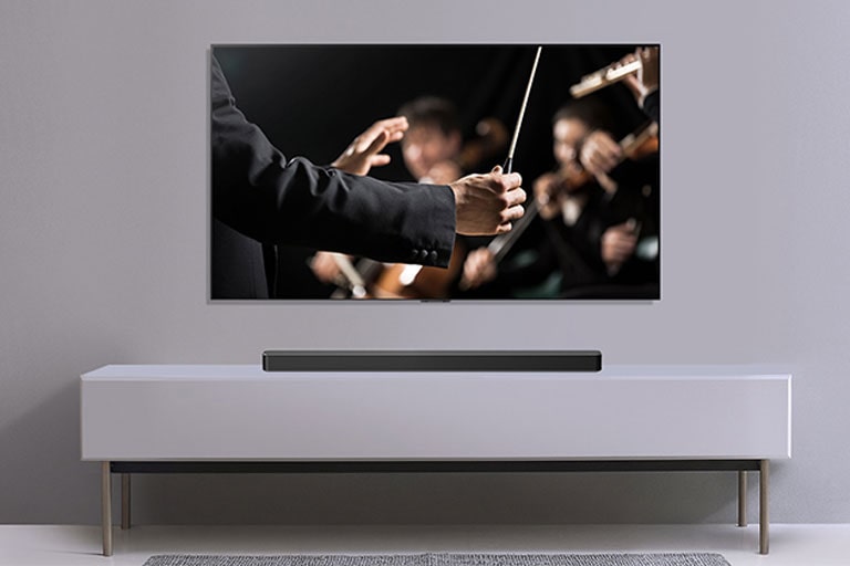 Un TV se muestra en una pared gris y la barra de sonido debajo de él en un mueble gris. El TV muestra a un director de orquesta.