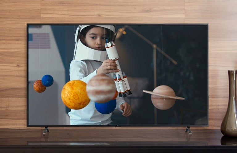 La pantalla de un televisor que reproduce un video de un niño con un traje de astronauta hecho por él, jugando con una nave espacial en su habitación decorada con miniaturas de planetas (reproducir el video)