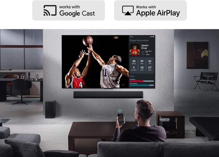 Una persona en su sala de estar sostiene su teléfono. En el teléfono, un icono de transmisión indica que la pantalla se está reflejando en el televisor. En el televisor, a un lado, se ve un partido de baloncesto y la pantalla reflejada muest las estadísticas de los jugadores.