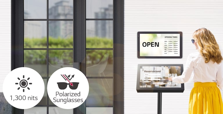 Hay una 22XF1TJ instalada sobre la pared en la entrada del edificio con otra 22XF1TJ debajo instalada sobre un pie para hacer reservaciones. Una mujer con lentes de sol está usando la pantalla táctil para hacer una reservación. La pantalla se puede ver incluso bajo la luz solar directa.
