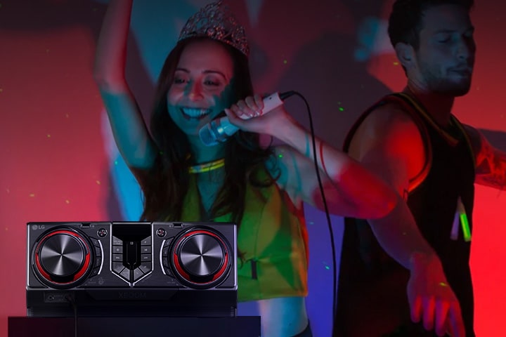 La imagen muestra una fiesta con luces de neón rojas. La gente disfruta de la música; uno de ellos sostiene un micrófono mientras los otros dos bailan. En el lado izquierdo de la imagen hay una mesa con un receptor colocado encima.