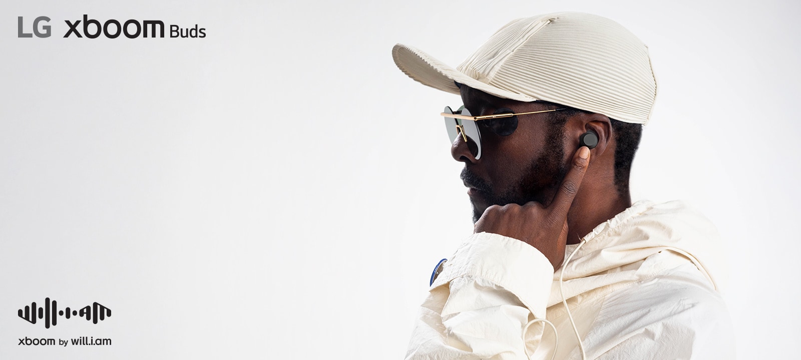 will.i.am con traje blanco y gafas de sol está mirando hacia el lado derecho apuntando el auricular a su oreja con su dedo índice izquierdo.