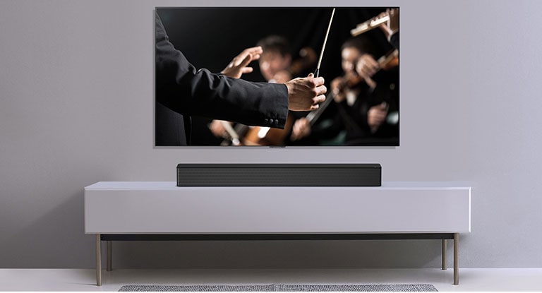 TV en una pared gris y barra de sonido LG abajo en un estante gris. La TV tiene un director dirigiendo una orquesta.