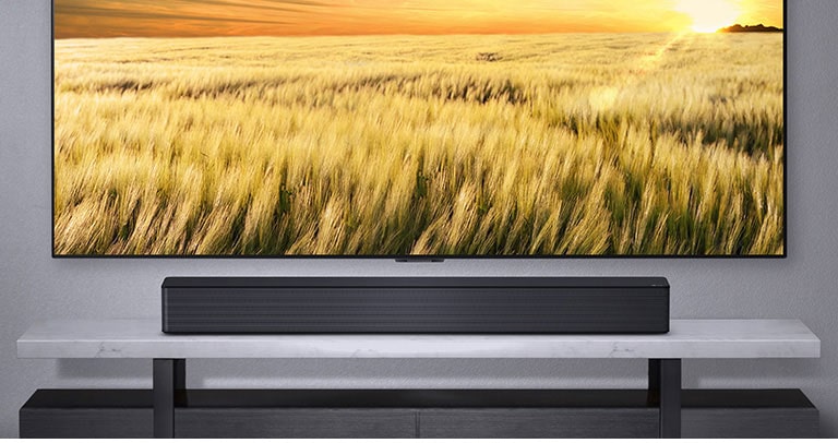 Se ve una TV en una pared gris y una barra de sonido LG debajo en un estante gris. Disco Blue-Ray debajo del estante.