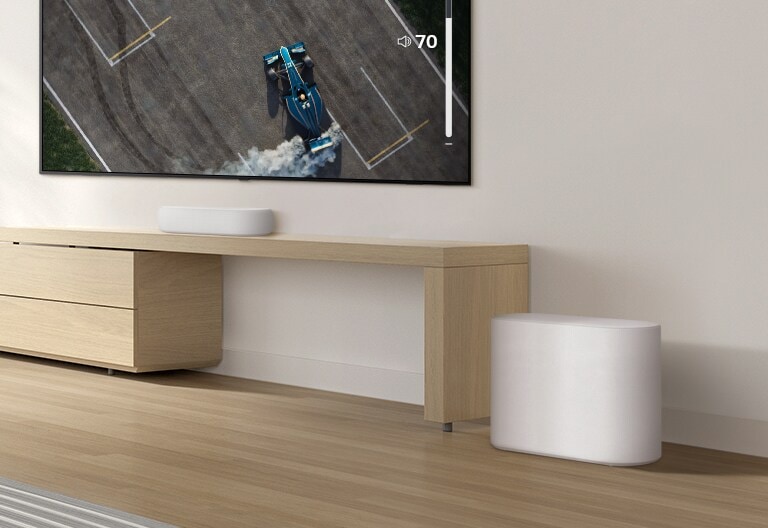 Una barra de sonido se coloca sobre una mesa de madera, un subwoofer se coloca en un piso de madera. Un suave efecto de onda de sonido sale del subwoofer en el suelo. Hay un televisor colocado encima de una barra de sonido. Un coche de carreras en la pantalla de un televisor se conduce muy rápido y violentamente y la GUI del volumen del televisor aumenta.