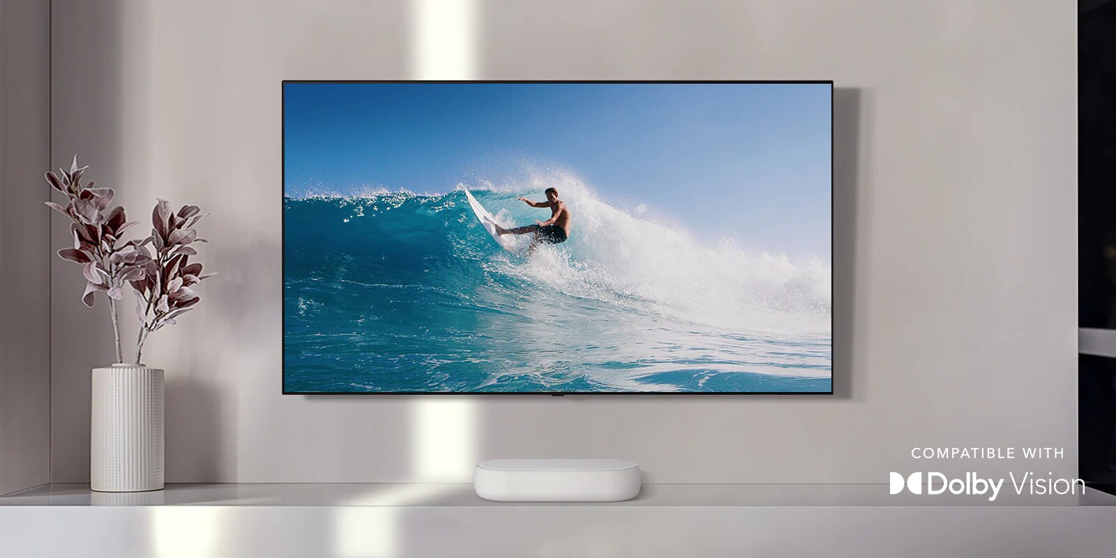 La televisión está en la pared. La televisión muestra a un hombre navegando en una gran ola. La barra de sonido LG está justo debajo del televisor en un estante blanco. Hay un jarrón con una flor justo al lado de la barra de sonido y el logotipo de Dolby Vision en la esquina inferior derecha.