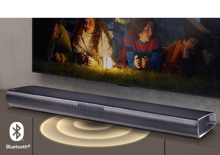 El televisor LG está en la pared, en la pantalla aparecen dos parejas tumbadas en la hierba. Delante de ellos hay una lámpara. La barra de sonido LG está debajo del televisor LG. El gráfico de sonido sale de la parte delantera de la barra de sonido. El logotipo de Bluetooth aparece en la esquina inferior izquierda de la imagen.