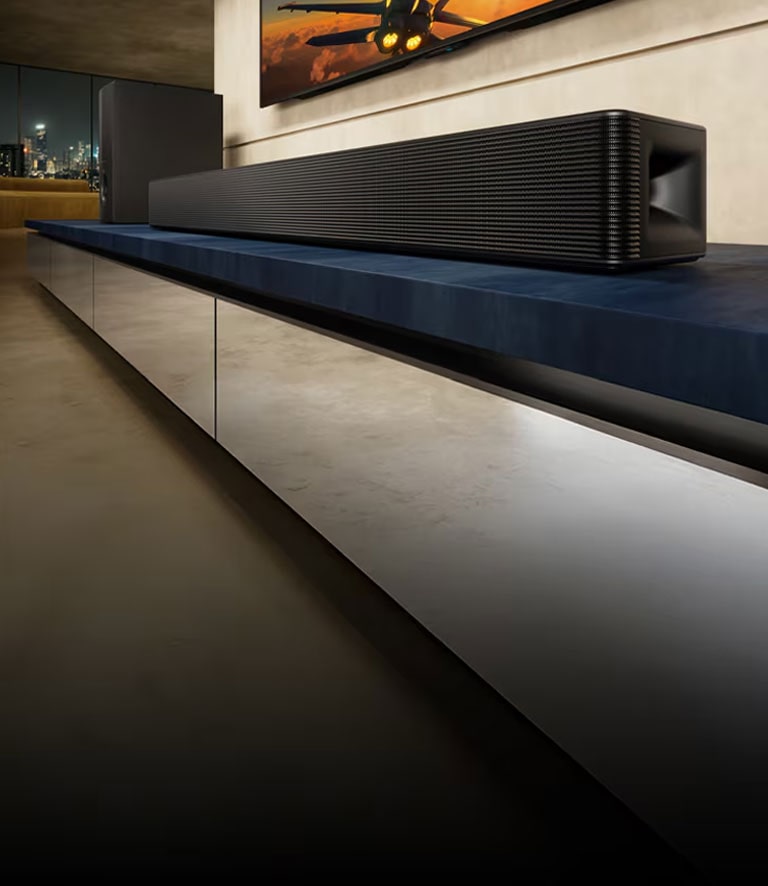 La barra de sonido LG SH5A y su subwoofer se encuentran sobre un mueble de televisión azul en una sala de estar. El televisor con la imagen de un avión se muestra parcialmente sobre la barra de sonido.