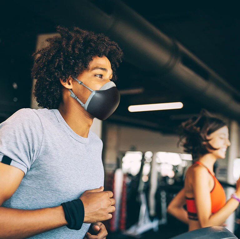 Un hombre afroamericano corre sobre unacaminadora dentro de un gimnasio y lleva puesto el purificador de aire facial LG PuriCare de color negro. Parece respirar sin agitarse.