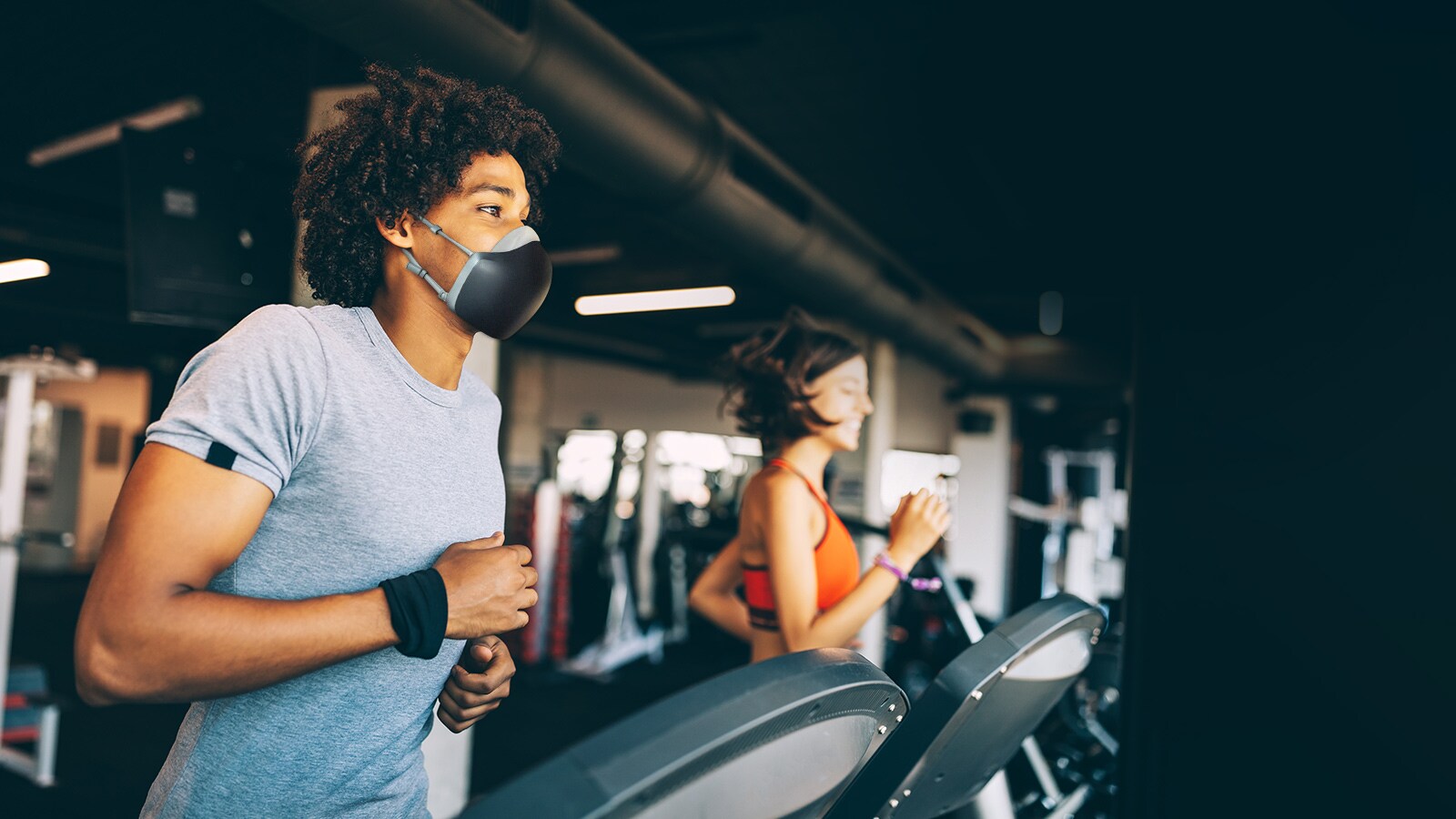 Un hombre afroamericano corre sobre una caminadora dentro de un gimnasio y lleva puesto el purificador de aire facial  LG PuriCare de color negro. Parece respirar sin agitarse.