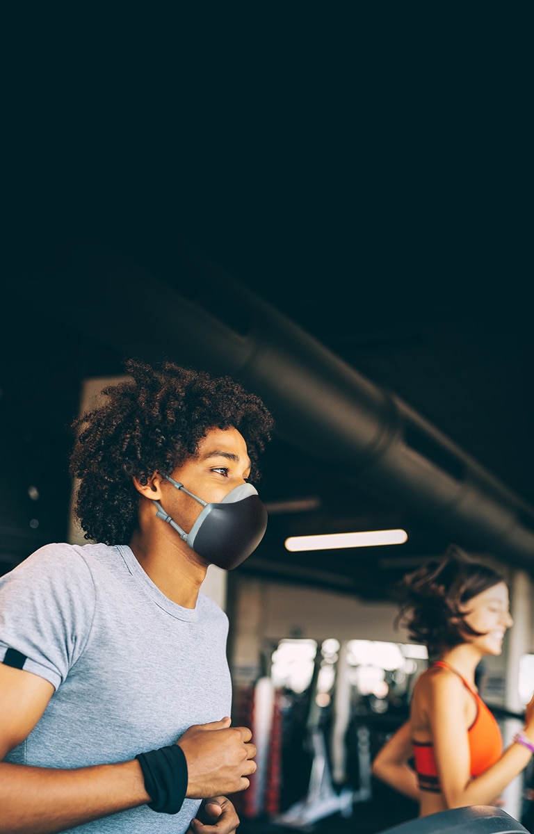 Un hombre afroamericano corre sobre una caminadora dentro de un gimnasio y lleva puesto el purificador de aire facial  LG PuriCare de color negro. Parece respirar sin agitarse.