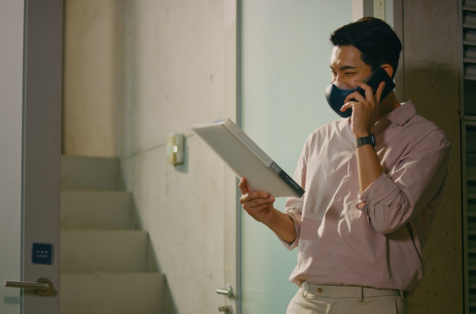 Un hombre asiático sostiene un documento y habla por teléfono. Lleva puesto un purificador de aire facial LG PuriCare™ negro.