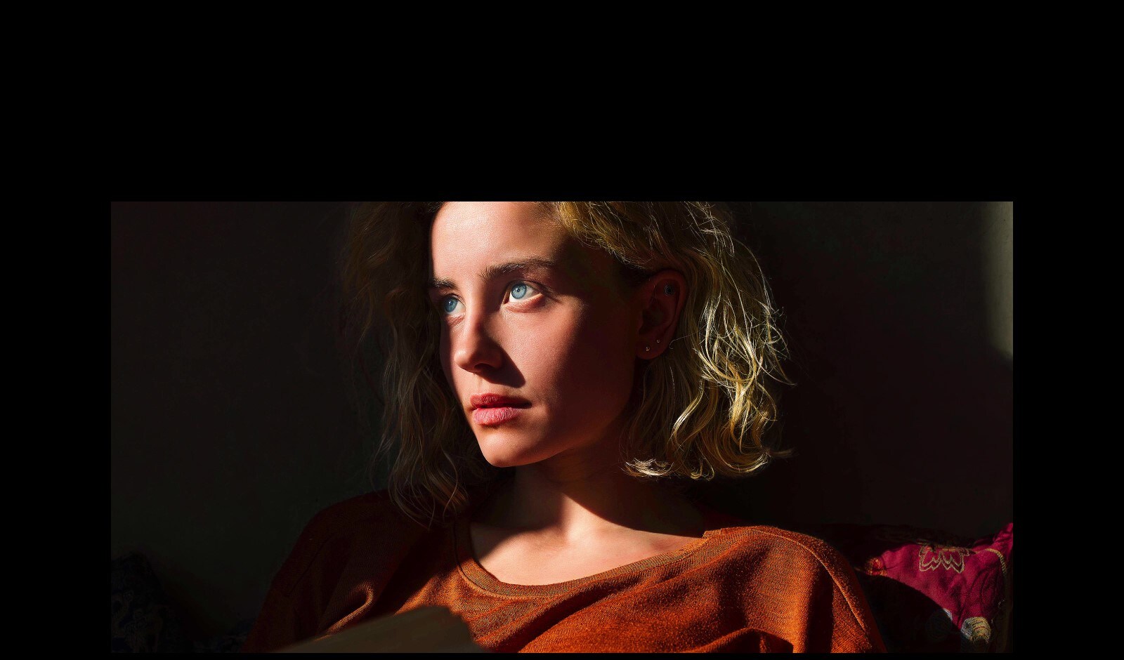 Una mujer con penetrantes ojos azules y un top naranja oscuro en un espacio oscuro. Las líneas rojas que representan los refinamientos de la IA cubren parte de su rostro, que es brillante y detallado, mientras que el resto de la imagen parece menos colorida.