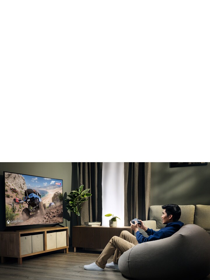Una persona sentada en un puf juega un juego de carreras en un televisor grande usando el controlador Bluetooth LG Razer Wolverine, con una planta visible cerca en una acogedora sala de estar.