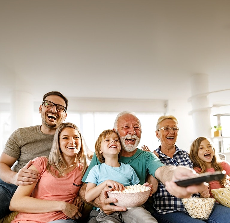 Una familia sentada riendo mientras el abuelo apunta con el control remoto.