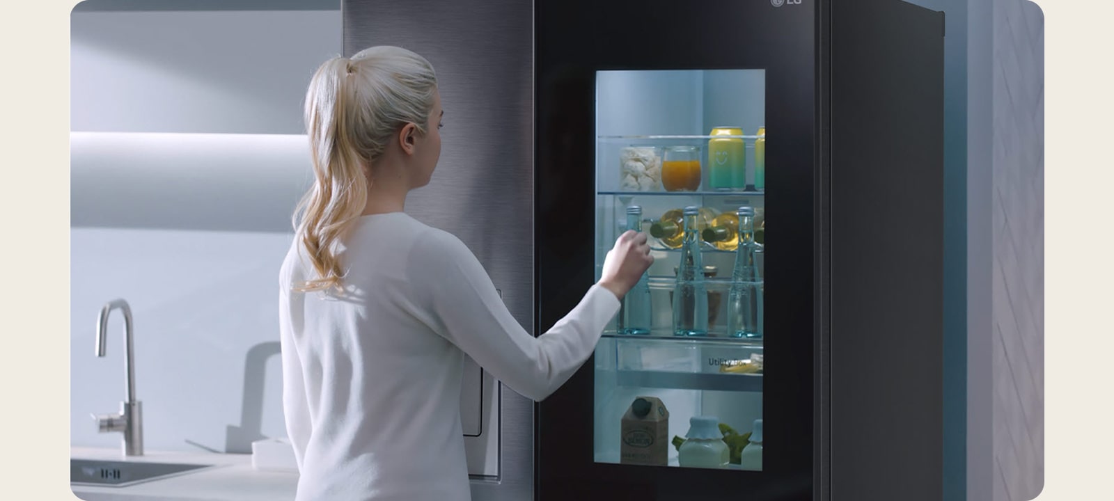 Un video muestra a una mujer acercándose a su refrigerador InstaView y tocando dos veces. El interior se ilumina y ella puede ver el contenido del refrigerador sin abrir la puerta. La vista hace un acercamiento para enfocarse en las bebidas de la puerta y luego se aleja para mostrar a la mujer desde atrás mientras abre la puerta y toma una bebida.