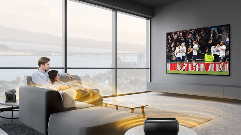 Un hombre y una mujer mirando deporte en el televisor del salón con unos altavoces Bluetooth traseros