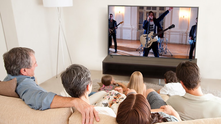 Una familia de siete personas reunida en la sala, viendo una película. La pantalla de TV muestra una banda tocando.