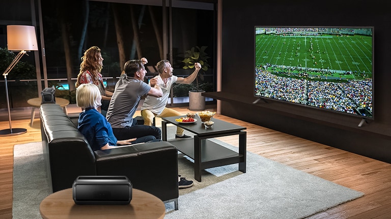 Personas que ven juegos deportivos en la TV en la sala de estar con altavoces traseros Bluetooth.