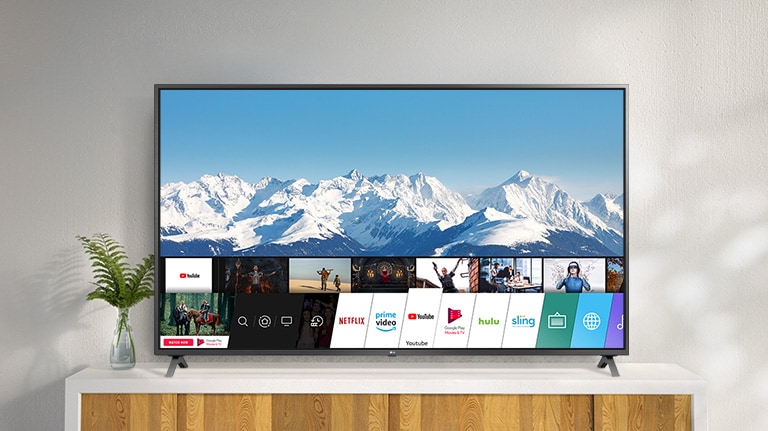 TV parada sobre un soporte blanco contra una pared blanca. La pantalla de TV muestra la pantalla de inicio con webOS.