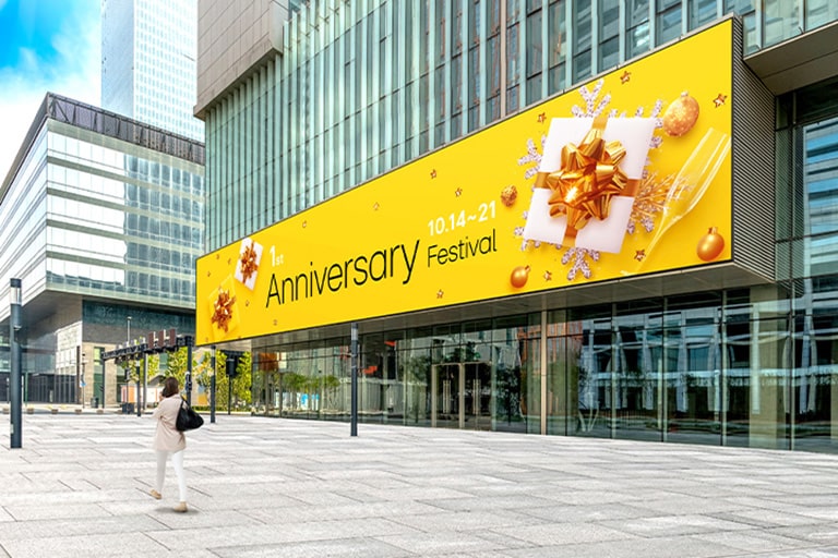 A large LED screen is installed on the exterior wall of the building entrance, and a woman is watching the advertisements on its screen. The screen remains clear and visible despite the bright sunlight.	